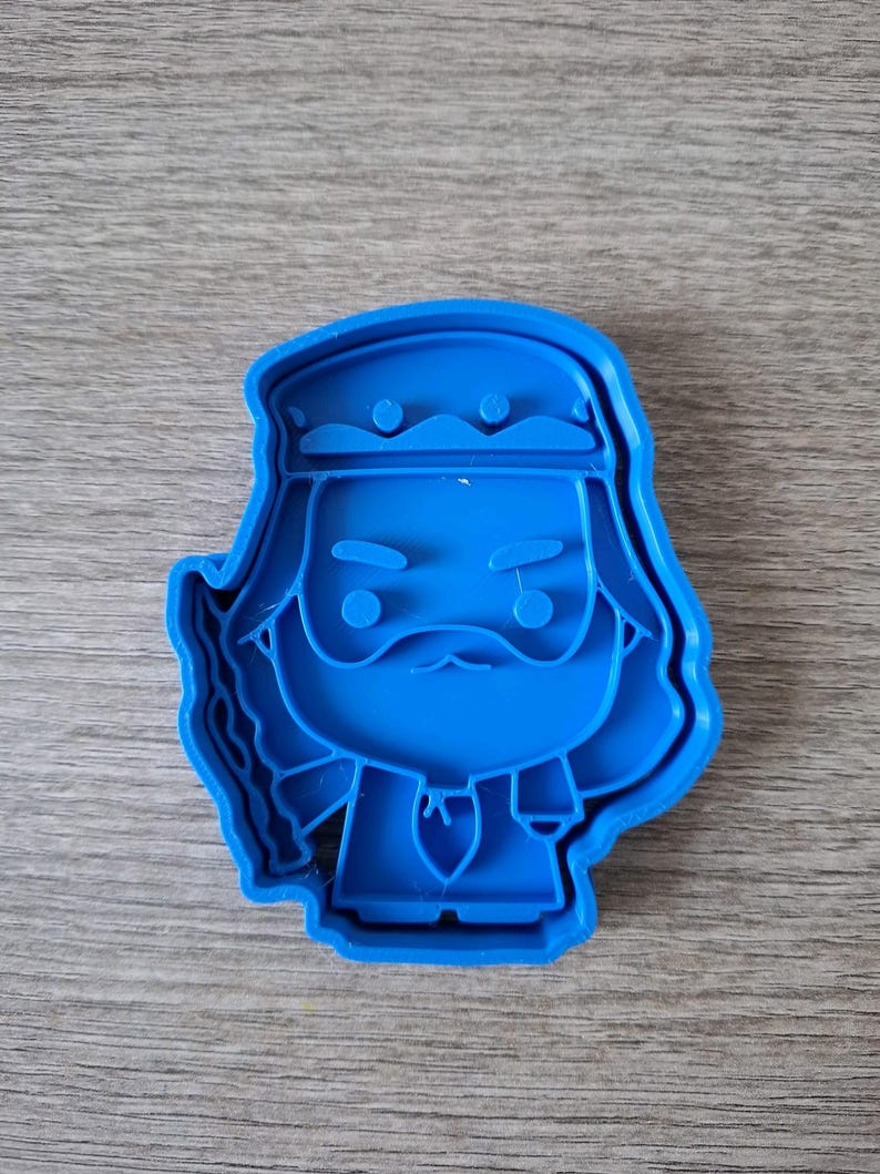 May include: A blue plastic cookie cutter in the shape of a cartoon character with a beard, mustache, and a head covering. The cutter has a detailed outline and is designed for creating cookies or other treats.