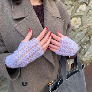 May include: Pale lilac fingerless gloves with a delicate openwork pattern. The gloves are made of a soft, knitted material. The person is wearing a grey coat and holding a grey tote bag.