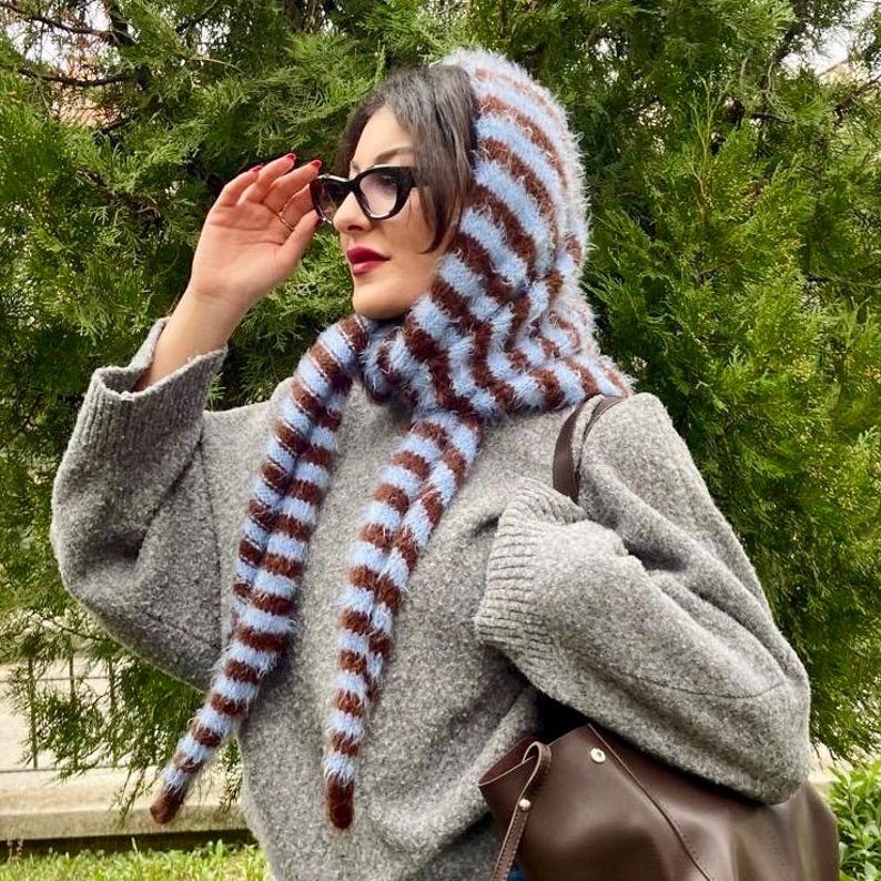 Puede incluir: Un su&eacute;ter gris con una capucha y bufanda a juego a rayas marrones y azul claro. La capucha y la bufanda son esponjosas y texturizadas. La persona lleva gafas negras y las sostiene con la mano.