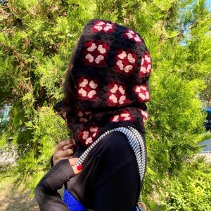 Könnte beinhalten: Eine schwarze Häkel-Balaclava mit einem floralen Muster in Rot und Hellrosa. Die Balaclava besteht aus weichem, strukturiertem Garn und bedeckt Kopf und Hals. Sie wird mit einem schwarzen Pullover und einer blauen Tasche getragen.