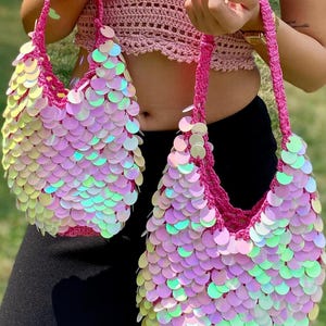 May include: Two matching crochet handbags featuring a vibrant pink yarn base covered in iridescent, circular sequins that reflect light with a multi-colored sheen. Each bag has a sturdy, braided pink handle and a rounded, slouchy silhouette.