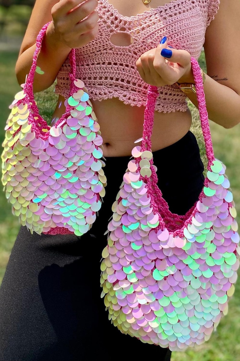 May include: Two matching crochet handbags featuring a vibrant pink yarn base covered in iridescent, circular sequins that reflect light with a multi-colored sheen. Each bag has a sturdy, braided pink handle and a rounded, slouchy silhouette.
