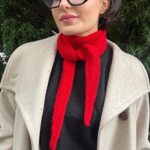 May include: A bright red knitted scarf is knotted at the neck, with long ends hanging down. The scarf is worn over a black sweater and a light beige coat. The background is a blurred view of green foliage.