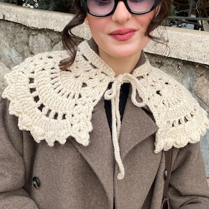 May include: A cream-colored crocheted collar featuring an intricate openwork pattern with a scalloped edge. The accessory is designed to be worn over clothing and includes two long crocheted ties at the center front for securing the piece in place.