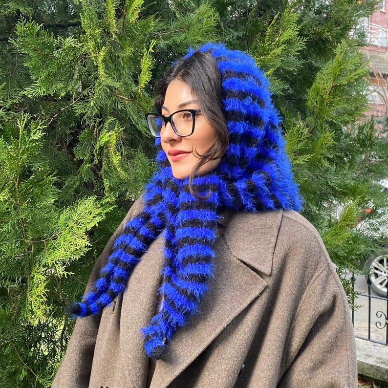 Puede incluir: Un conjunto de capucha y bufanda a rayas azules y negras. La capucha y la bufanda est&aacute;n hechas de un material esponjoso y texturizado. La bufanda cuelga por delante de un abrigo marr&oacute;n. La persona lleva gafas negras.