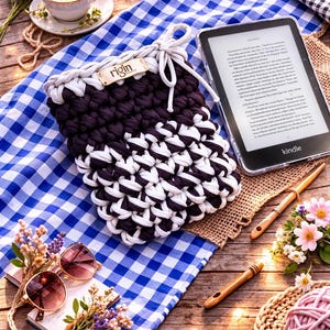May include: A handmade crochet Kindle sleeve in black and white, with a white drawstring closure. The sleeve rests on a blue and white checkered cloth, alongside a Kindle e-reader, sunglasses, and flowers.