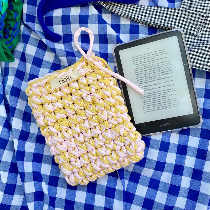 May include: A handmade crochet Kindle sleeve in yellow and pink. The sleeve has a loop handle and the brand name "rigin" is visible. A Kindle e-reader is next to the sleeve on a blue and white checkered surface.
