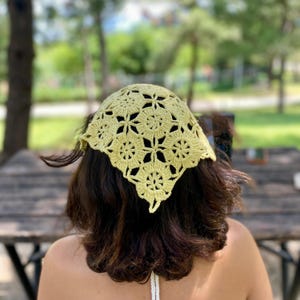 May include: A light yellow crocheted headscarf with a floral pattern. The head covering is square-shaped and worn on the head. The background is blurred, showing a park setting with trees and a wooden table.