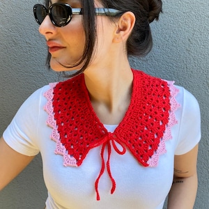 May include: A red and pink crochet collar with a tie closure. The collar is a vibrant red with a scalloped pink trim. The collar is worn over a white t-shirt.