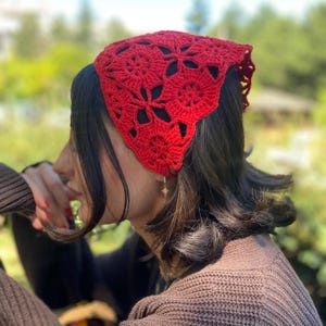 May include: A vibrant red crochet headscarf featuring a floral pattern. The head covering is made of a soft, textured yarn and is worn on the head. The headscarf is a stylish accessory.