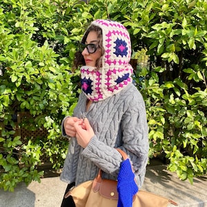 Può includere: Un set di cappuccio e sciarpa all'uncinetto color crema con un motivo geometrico rosa e blu navy. Il cappuccio e la sciarpa sono indossati sopra un maglione grigio a trecce. È visibile anche una borsa beige con dettagli in pelle marrone e una sciarpa blu.