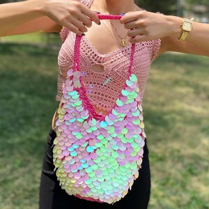May include: A crochet shoulder bag featuring a vibrant pink yarn base completely covered in iridescent, circular, scale-like sequins that reflect light in shades of green, blue, and pink. The bag has a single, thick, braided crochet strap.