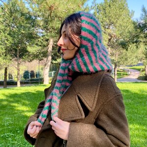 Puede incluir: Una persona con una capucha y bufanda de punto a rayas rosas y verdes. La bufanda est&aacute; envuelta alrededor del cuello y la capucha cubre la cabeza. La persona lleva un abrigo marr&oacute;n y est&aacute; en un parque con hierba verde y &aacute;rboles.