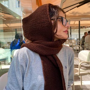 Könnte beinhalten: Ein dunkelbrauner, gestrickter Kapuzenschal mit texturiertem Kraus-rechts-Muster. Der Schal verfügt über eine integrierte Kapuze, die den Kopf bedeckt und nach vorne fällt, getragen über einem hellgrauen Langarmpullover mit kleinem Sticklogo.