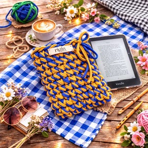 May include: A handmade blue and yellow knitted Kindle sleeve with a drawstring closure. The sleeve is placed on a blue and white checkered cloth, next to a Kindle e-reader, a cup of coffee, and a pair of sunglasses.