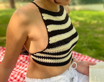 May include: A black and cream-colored striped crochet halter top. The top is handmade with a halter neck and a cropped design. The person is wearing white pants and sunglasses while sitting on a red and white checkered blanket.