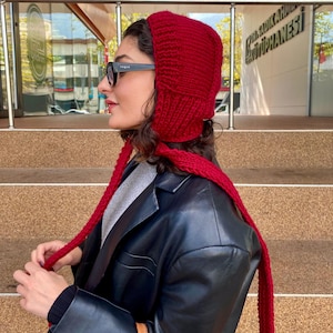 May include: A knitted red hood with long ties. The hood covers the head and neck, with the ties hanging down. The person in the image is wearing black sunglasses and a black leather jacket.