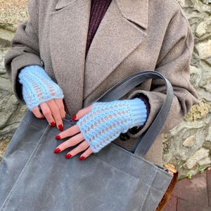 May include: Pale blue fingerless gloves with a detailed openwork pattern. The gloves are worn with a brown coat and a grey tote bag. The nails are painted red.