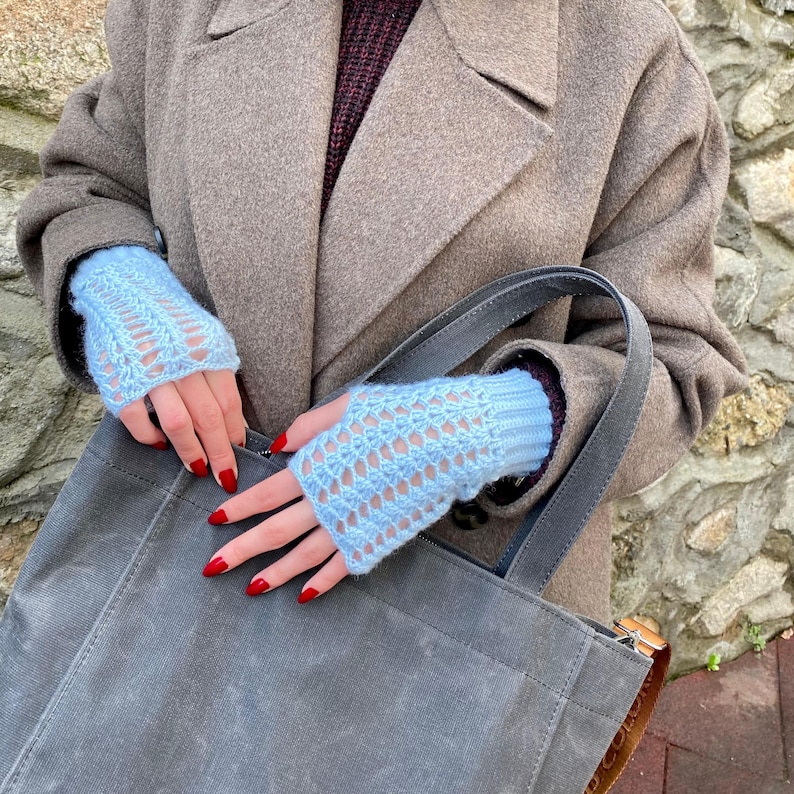May include: Pale blue fingerless gloves with a detailed openwork pattern. The gloves are worn with a brown coat and a grey tote bag. The nails are painted red.