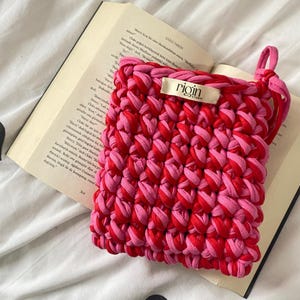 May include: A handmade, square crochet bag in shades of red and pink, with a loop handle. The bag is resting on an open book. The label on the bag reads "rigin studio".