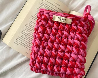 May include: A handmade, square crochet bag in shades of red and pink, with a loop handle. The bag is resting on an open book. The label on the bag reads "rigin studio".