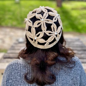 May include: A beige crocheted hat with a floral design and pearl accents. The hat is worn by a person with dark hair and a gray sweater. The background is blurred, showing a wooden surface and greenery.