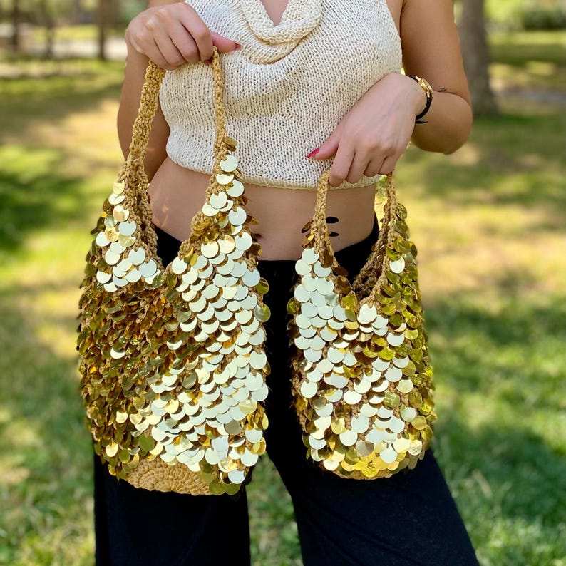 May include: Two gold crochet bags featuring a dense, overlapping pattern of shiny, circular metallic sequins. Each bag has a matching gold crocheted handle and a textured, woven base, creating a reflective, scale-like appearance on the exterior.