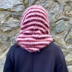 Puede incluir: Una capucha de punto en tonos rosa y burdeos, con rayas horizontales. La capucha est&aacute; hecha de un hilo difuso y se usa sobre una prenda de color oscuro. El fondo es una pared de piedra.