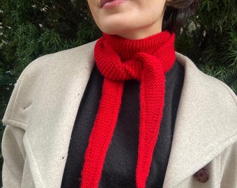 May include: A bright red knitted scarf tied in a knot around the neck, worn over a black sweater and a beige coat. The scarf features a textured garter stitch pattern with two long, pointed ends hanging down the front of the chest.