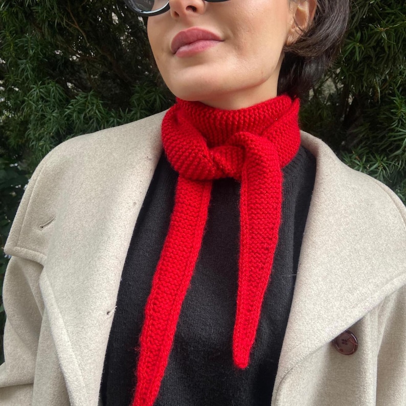 May include: A bright red knitted scarf is tied around the neck. The scarf is made of a thick yarn and has a textured appearance. The scarf is worn over a black sweater and a light beige coat.