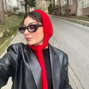 May include: A woman wearing a red knit hat and matching scarf with a black leather jacket and black cat-eye glasses.