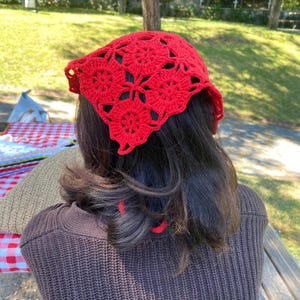 May include: A bright red, crocheted headscarf with a floral pattern. The head covering is made of a soft, open-weave material. The headscarf is worn on the head and covers the top of the head and the forehead.