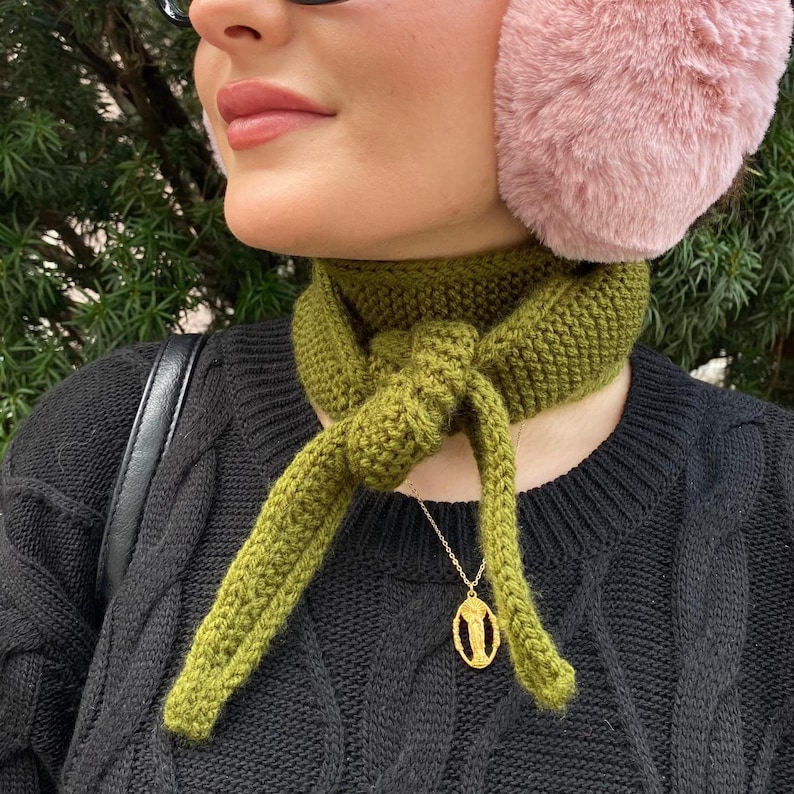 May include: A close-up of a person wearing a green knitted scarf tied in a knot. The person is also wearing a black sweater, pink earmuffs, and a gold pendant necklace. The background is blurred with green foliage.