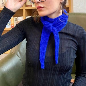 May include: A vibrant blue knitted scarf is knotted around the neck. The scarf is made of a thick yarn and hangs down in two long strands. The person is wearing a black long-sleeved shirt. The background includes a bookshelf and a green chair.