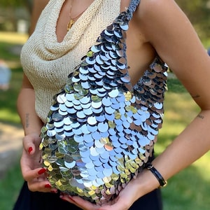 Può includere: Una borsa a sacco ricoperta di grandi paillettes circolari argentate e riflettenti. La borsa presenta una base in uncinetto grigio scuro e un unico tracolla integrata, creando un aspetto strutturato e scintillante con un motivo a scaglie.