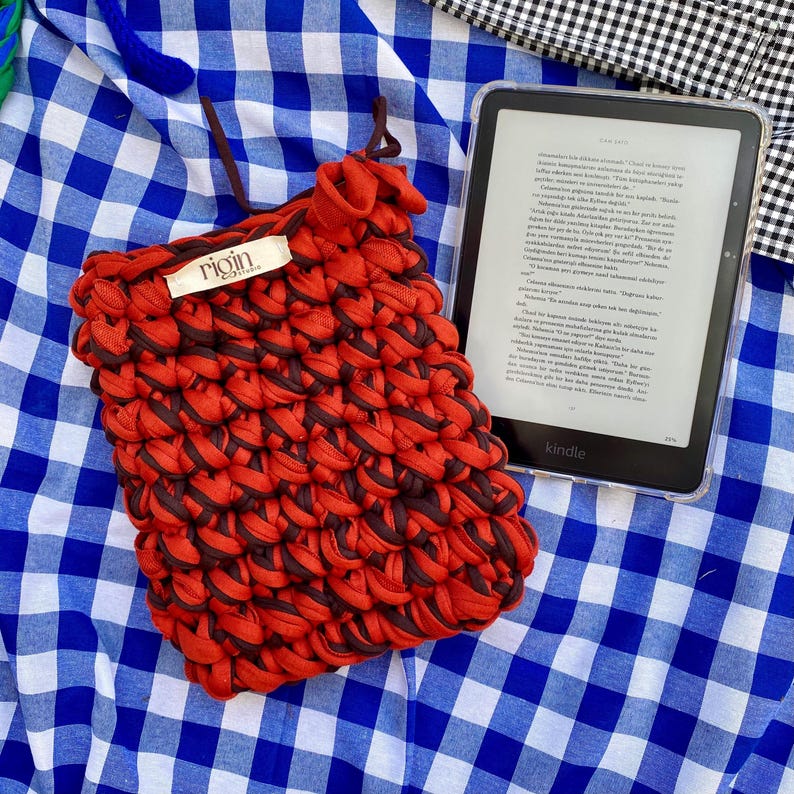 May include: A handmade red and brown woven Kindle sleeve with a drawstring closure. The sleeve features a textured design and a small label that reads "rigin". A Kindle e-reader is visible in the background, resting on a blue and white checkered surface.