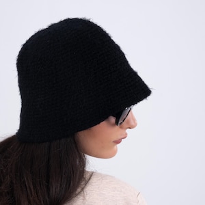 May include: A solid black bucket hat with a fuzzy, textured knit construction. The hat features a rounded crown and a downward-sloping brim that provides full coverage. The material appears soft and slightly fibrous, creating a matte finish.