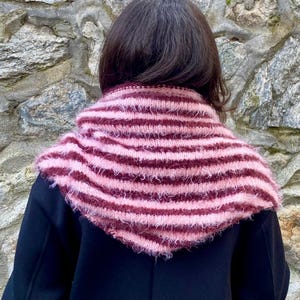 Puede incluir: Bufanda tubular de punto a rayas rosas y burdeos. La bufanda se lleva alrededor del cuello y los hombros, con una textura difusa. La persona lleva un abrigo negro. El fondo es una pared de piedra.