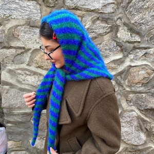 Puede incluir: Una capucha a rayas azules y verdes con extremos largos tipo bufanda. La capucha est&aacute; hecha de un material difuso y se usa sobre un abrigo marr&oacute;n. Los colores de la capucha son vibrantes y llamativos.