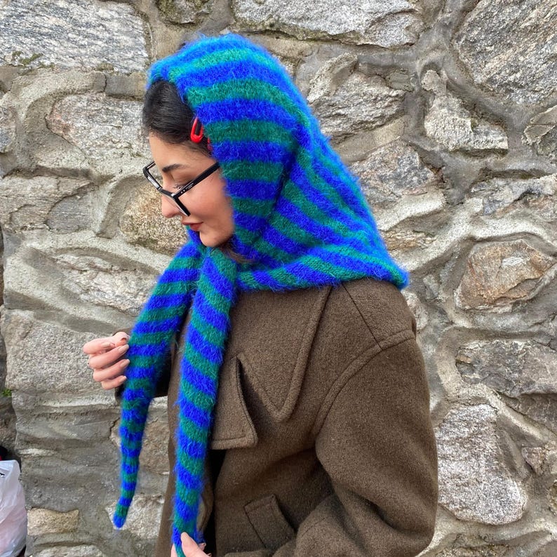 Puede incluir: Una capucha a rayas azules y verdes con extremos largos tipo bufanda. La capucha est&aacute; hecha de un material difuso y se usa sobre un abrigo marr&oacute;n. Los colores de la capucha son vibrantes y llamativos.