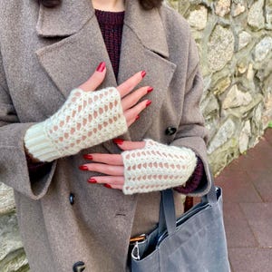May include: Cream-coloured, open-weave fingerless gloves. The gloves have a ribbed cuff and a delicate, diamond-shaped pattern across the hand. The gloves are worn with a brown coat and a grey tote bag.