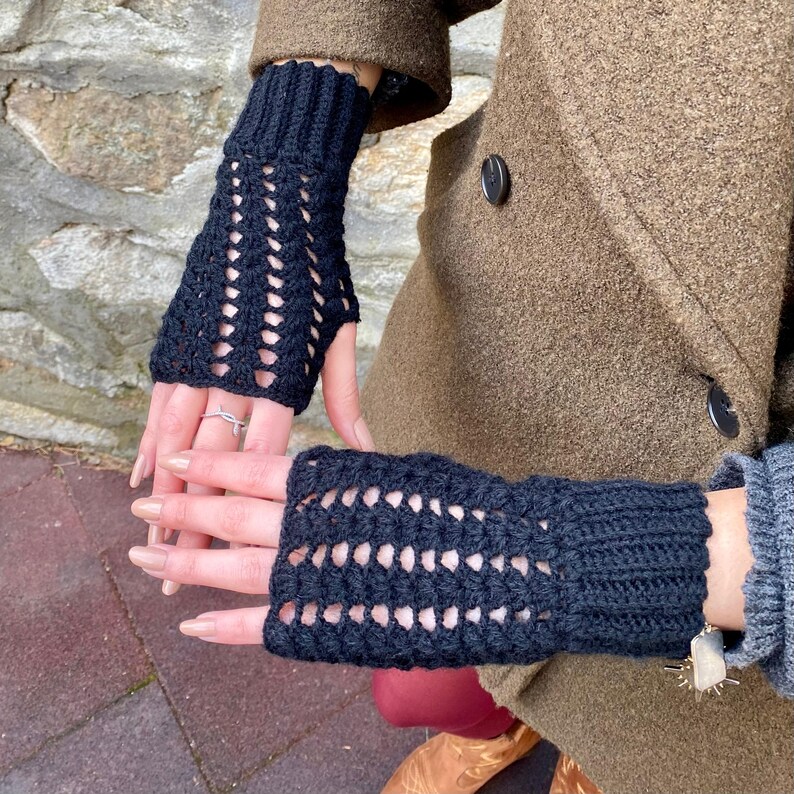May include: A pair of dark navy blue fingerless gloves. The gloves are made of a crocheted material with a repeating openwork pattern. The gloves are worn with a brown coat.