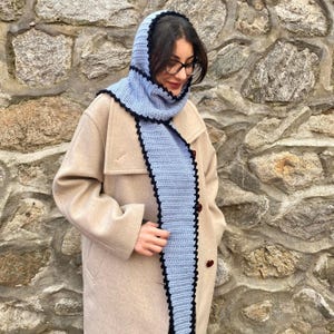 Può includere: Un set di cappuccio e sciarpa all'uncinetto blu chiaro con rifiniture nere. La sciarpa è lunga e rettangolare, e il cappuccio incornicia il viso. Il set è indossato sopra un cappotto beige.