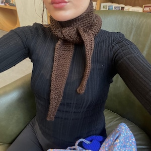 May include: A brown knitted scarf with long, dangling ends. The scarf is wrapped around the neck and hangs down the front. The scarf is made of a thick, textured yarn and is a solid brown color. The person is wearing a black long-sleeved shirt.