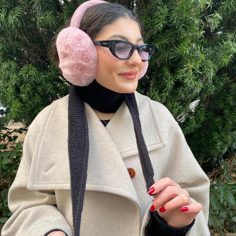 May include: A person wearing pink faux fur earmuffs and black-framed glasses. The person is also wearing a black scarf and a light beige coat. The earmuffs are fluffy and cover the ears. The glasses have a dark frame. The coat has a large collar.