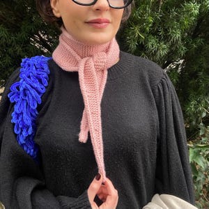 May include: A light pink knitted scarf tied in a knot. The scarf is made of a soft, textured yarn and is worn over a black sweater. A blue, fluffy shoulder bag is visible on the left side of the image.