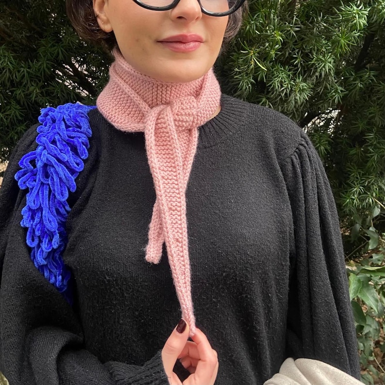 May include: A light pink knitted scarf tied in a knot. The scarf is made of a soft, textured yarn and is worn over a black sweater. A blue, fluffy shoulder bag is visible on the left side of the image.