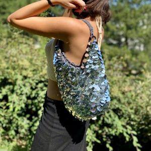 May include: A hobo-style shoulder bag crafted from grey crocheted yarn and covered in large, reflective, circular silver sequins that overlap like scales. The bag features a single wide strap and a slouchy, unstructured silhouette.