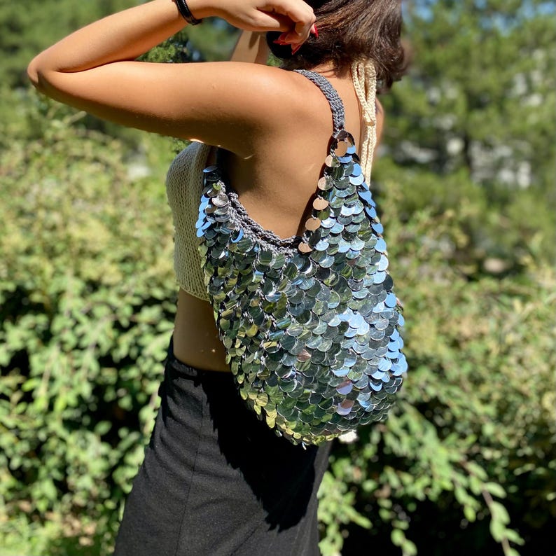 May include: A hobo-style shoulder bag crafted from grey crocheted yarn and covered in large, reflective, circular silver sequins that overlap like scales. The bag features a single wide strap and a slouchy, unstructured silhouette.