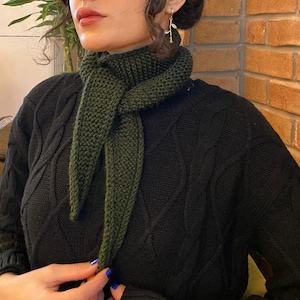 May include: A dark green knitted scarf is draped around the neck, with the ends hanging down. The scarf is paired with a black sweater. The image is a close-up shot, highlighting the texture and color of the scarf.